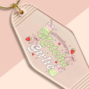 Peut inclure: Un porte-clés rose avec un anneau doré. Le porte-clés affiche les mots "Matcha Girlie" dans une police fantaisiste, avec des illustrations de fraises et de boissons. Le design comprend des couleurs rose et vert.