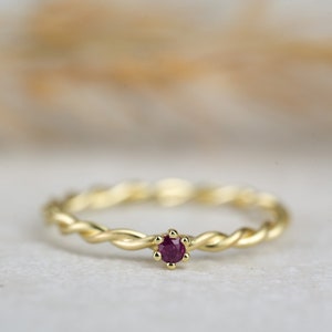 Könnte beinhalten: Ein zarter Goldring mit einem gedrehten Design und einem kleinen, runden, dunkelroten Edelstein in der Mitte.