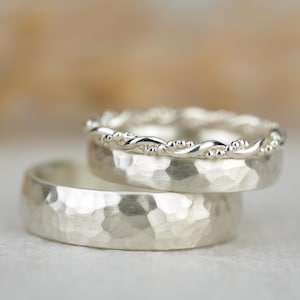 Op de afbeelding: Twee zilveren ringen op elkaar gestapeld. De onderste ring heeft een gehamerde textuur, terwijl de bovenste ring een gedraaid touwontwerp heeft. Waarschijnlijk trouwringen of belofteringen.