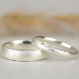 Könnte beinhalten: Zwei silberne Eheringe mit gebürsteter Oberfläche. Die Ringe liegen übereinander.
