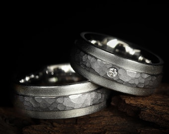 May include: Two silver wedding rings with a textured hammered finish. One ring features a small diamond. The rings are displayed on a dark wooden surface against a black background. The rings are likely made of titanium or a similar metal.