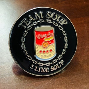 Könnte beinhalten: Schwarzer Emaille-Pin mit silbernem Kettenrand. Der Pin zeigt eine rote und weiße Suppendose mit dem Text "Whop Ass" und "Soup". Der Text um die Dose lautet "Team Soup I Like Soup".