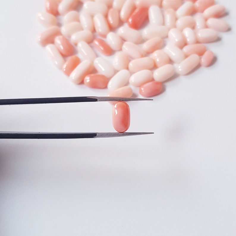 May include: A close-up of a single, oval-shaped, peach-colored gemstone held by tweezers. The gemstone is surrounded by a scattering of similar, smaller gemstones in varying shades of peach and white, all on a white background.