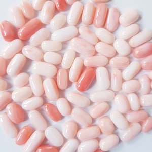 May include: A collection of oval-shaped coral beads in varying shades of pink and white. The beads are smooth and polished, with a glossy finish. The colors range from pale blush to a deeper, more saturated coral hue. The beads are scattered across a white surface.
