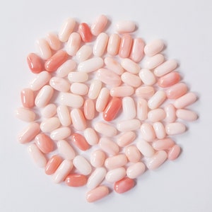 May include: A collection of oval-shaped coral beads in varying shades of pink and white. The beads are smooth and polished, with some displaying a deeper, more vibrant pink hue. The arrangement is scattered on a white surface.