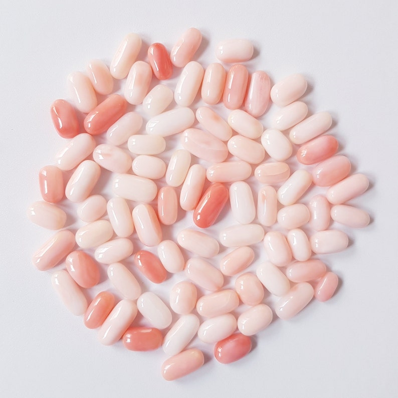 May include: A collection of oval-shaped coral beads in varying shades of pink and white. The beads are smooth and polished, with some displaying a deeper, more vibrant pink hue. The arrangement is scattered on a white surface.