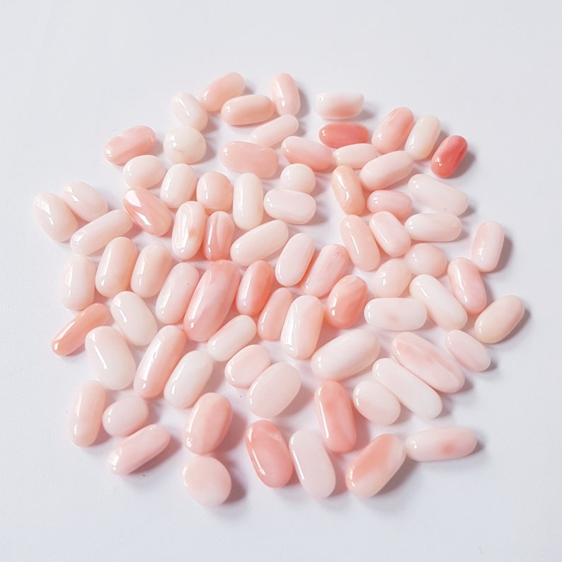 May include: A collection of oval-shaped, polished gemstones in varying shades of pink and peach. The gemstones are scattered on a white surface, with some displaying a translucent quality.