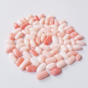 May include: A collection of oval-shaped coral beads in varying shades of pink and white. The beads are smooth and polished, with some displaying a deeper, more vibrant coral hue. The arrangement is scattered, creating a natural and organic aesthetic.