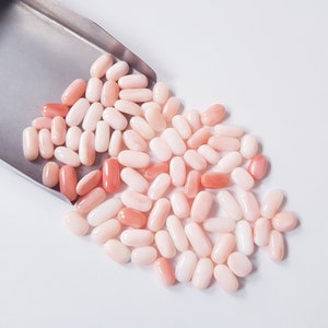 May include: A collection of oval-shaped coral beads in varying shades of pink and peach, spilling from a silver scoop onto a white surface. The beads are smooth and polished, showcasing natural color variations.