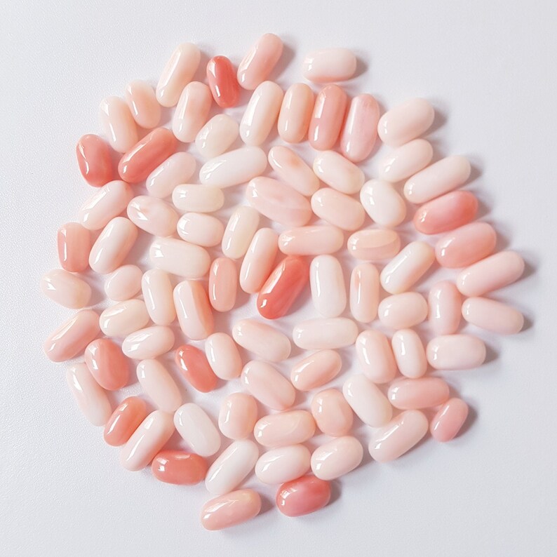 May include: A collection of oval-shaped coral beads in varying shades of pink and peach. The beads are smooth and polished, with a natural, organic appearance. The arrangement is scattered, showcasing the color variations.