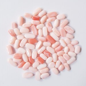 May include: A collection of oval-shaped coral beads in varying shades of pink and peach. The beads are smooth and polished, with a natural, organic appearance. The beads are scattered on a white surface.