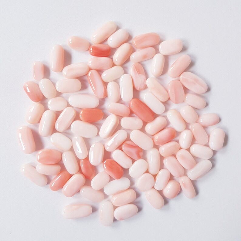 May include: A collection of oval-shaped coral beads in varying shades of pink and peach. The beads are smooth and polished, with a natural, organic appearance. The beads are scattered on a white surface.