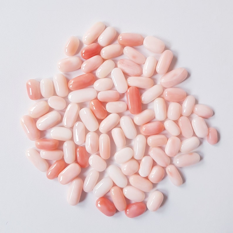 May include: A collection of oval-shaped coral beads in varying shades of pink and peach. The beads are smooth and polished, with a natural, organic appearance. They are scattered on a white surface.