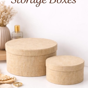Könnte beinhalten: Zwei runde Jute-Aufbewahrungsboxen in verschiedenen Größen. Die Boxen sind beige und haben eine strukturierte Oberfläche. Der Text "Round Jute Storage Boxes" befindet sich oben im Bild.