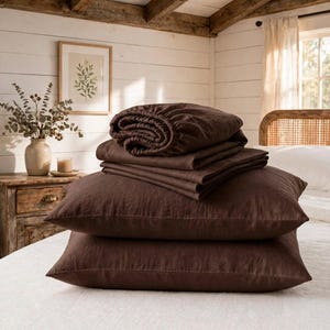 May include: A set of dark brown bedding featuring two rectangular pillows stacked at the base, topped with a folded flat sheet and a fitted sheet. The fabric has a visible woven texture, and the items are arranged neatly on a white surface.