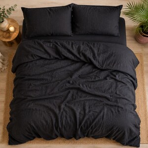 May include: A charcoal black bedding set consisting of a duvet cover and two matching rectangular pillows. The fabric has a visible textured weave, and the duvet cover is draped over the bed with soft folds and creases across the surface.