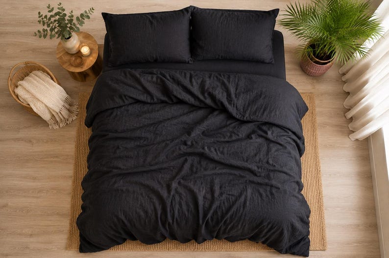 May include: A charcoal black bedding set consisting of a duvet cover and two matching rectangular pillows. The fabric has a visible textured weave, and the duvet cover is draped over the bed with soft folds and creases across the surface.