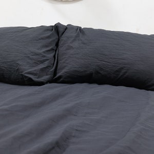 May include: A dark charcoal gray bedding set consisting of a duvet cover and two matching rectangular pillows. The fabric has a soft, slightly wrinkled texture and a matte finish, covering the entire surface of the bed in a uniform dark tone.