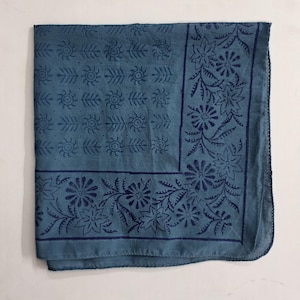 May include: A folded teal-colored bandana with a dark blue floral and leaf pattern. The design includes repeating sun-like shapes and floral borders. The fabric appears to be cotton or a similar material.
