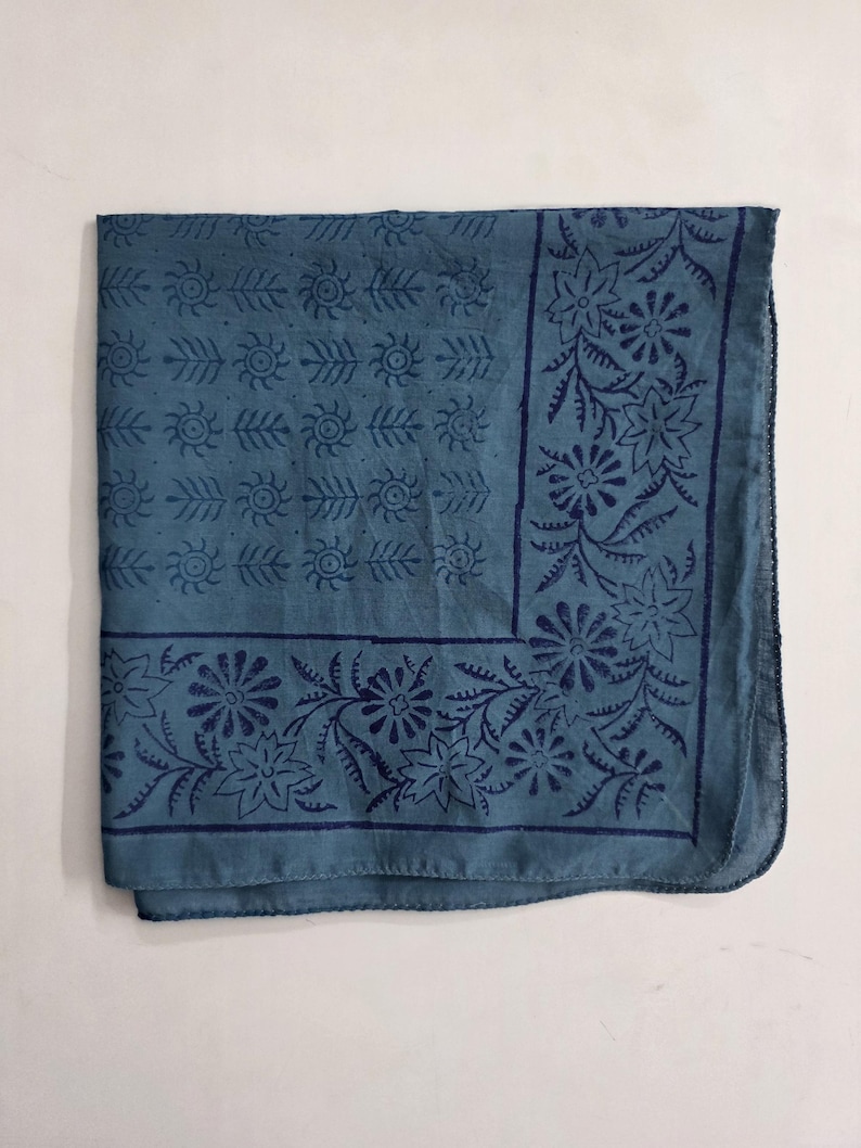 May include: A folded teal-colored bandana with a dark blue floral and leaf pattern. The design includes repeating sun-like shapes and floral borders. The fabric appears to be cotton or a similar material.