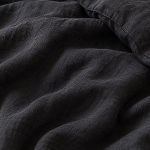 May include: A dark charcoal gray textile with a visible woven texture and soft folds. The fabric displays a matte finish and a slightly crinkled appearance, with a straight stitched seam visible along the upper right edge of the material.
