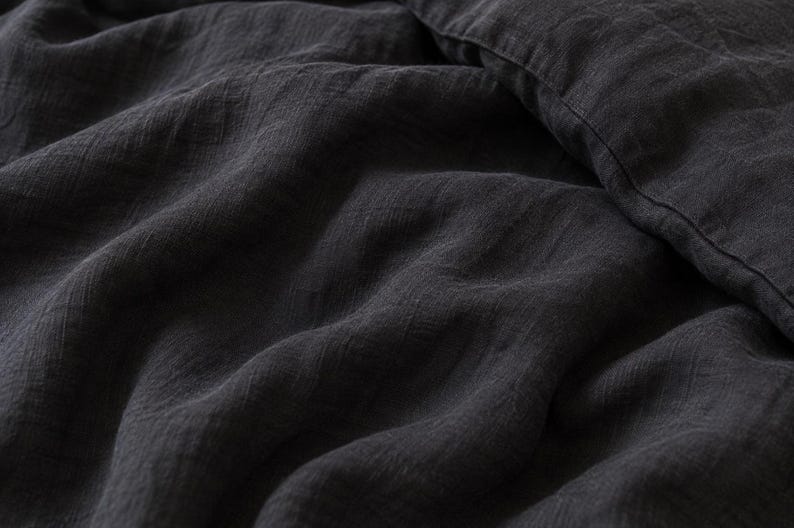 May include: A dark charcoal gray textile with a visible woven texture and soft folds. The fabric displays a matte finish and a slightly crinkled appearance, with a straight stitched seam visible along the upper right edge of the material.