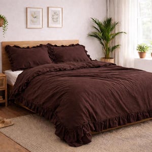 May include: A bedroom scene featuring a bed with a dark brown ruffled duvet cover and matching pillowcases. A wooden nightstand with a lamp and decorative items sits beside the bed. The room has a neutral color palette with natural light.