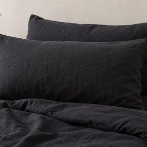 May include: A set of charcoal black rectangular pillowcases and a matching duvet cover made of a textured fabric. The bedding features a visible weave and a matte finish, arranged neatly on a bed with multiple pillows stacked against each other.