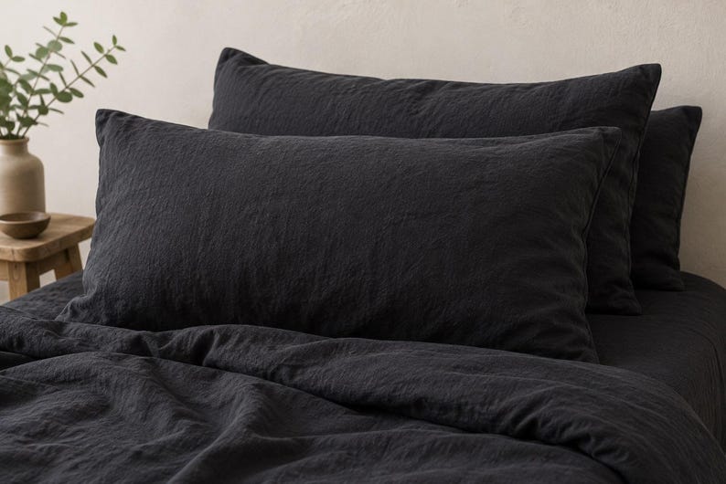 May include: A set of charcoal black rectangular pillowcases and a matching duvet cover made of a textured fabric. The bedding features a visible weave and a matte finish, arranged neatly on a bed with multiple pillows stacked against each other.