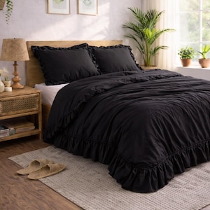 May include: A bedroom scene featuring a bed with a black ruffled duvet cover and matching pillows. A wooden nightstand with a lamp and a woven rug are in the foreground. Two framed botanical prints hang on the wall.