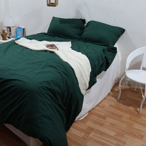 Può includere: Un copripiumino verde su un letto con cuscini bianchi e una coperta bianca. Una sedia in vimini bianca si trova nell'angolo della stanza.