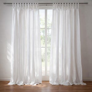 May include: White linen curtains hanging from a silver rod, framing a window with natural light. The curtains are long, with a slightly textured appearance, and are gathered at the top with fabric loops. The floor is a warm brown color.