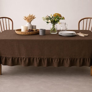 Op de afbeelding: Een bruin rechthoekig tafelkleed met een gerimpelde rand bedekt een houten tafel. De tafel is gedekt met witte borden, een vaas met bloemen en een gevlochten dienblad met kaarsen. Twee houten stoelen staan achter de tafel.