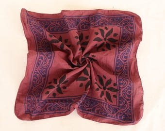 Könnte beinhalten: Ein weinrotes quadratisches Bandana mit dunkelblauem Paisley-Rand und schwarzem floralem Mitteldesign. Der Stoff ist spiralförmig gefaltet, wodurch die komplizierten Details des Drucks zur Geltung kommen. Dieses Accessoire ist vielseitig einsetzbar.