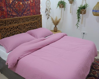 May include: A bed with a pink duvet cover and matching pillowcases. The bed has a carved wooden headboard and is set against a white wall. Decorative elements include hanging plants, a tapestry, and a rug.