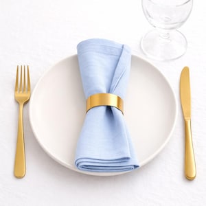 May include: A formal table setting featuring a white plate, gold fork and knife, and a light blue napkin held by a gold napkin ring. A wine glass is visible in the background. The setting is on a white surface.