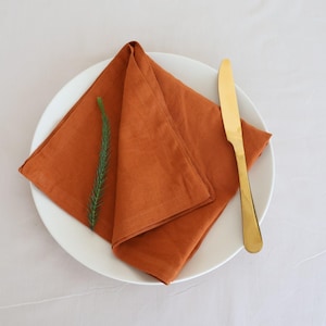 Könnte beinhalten: Eine gefaltete orangefarbene Leinen-Serviette auf einem weißen Teller mit einem goldenen Messer.