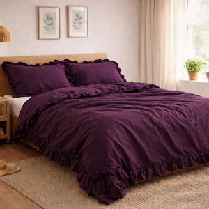 May include: A bedroom with a bed dressed in a deep purple duvet cover and pillowcases with a ruffled edge. The bed has a wooden headboard. A woven bedside table, botanical prints, and a woven rug create a warm and inviting atmosphere.