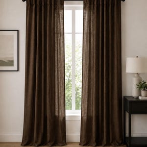May include: A pair of floor-length, dark brown curtains with a visible woven texture, hanging from a black curtain rod. The curtains are pleated at the top and drape vertically, framing a central window that allows natural light to pass through.