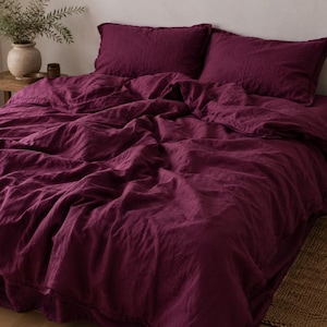 May include: A deep plum-colored bedding set featuring a duvet cover and two matching rectangular pillowcases. The fabric has a visible, slightly textured weave and is draped loosely over a bed, creating soft folds across the surface.