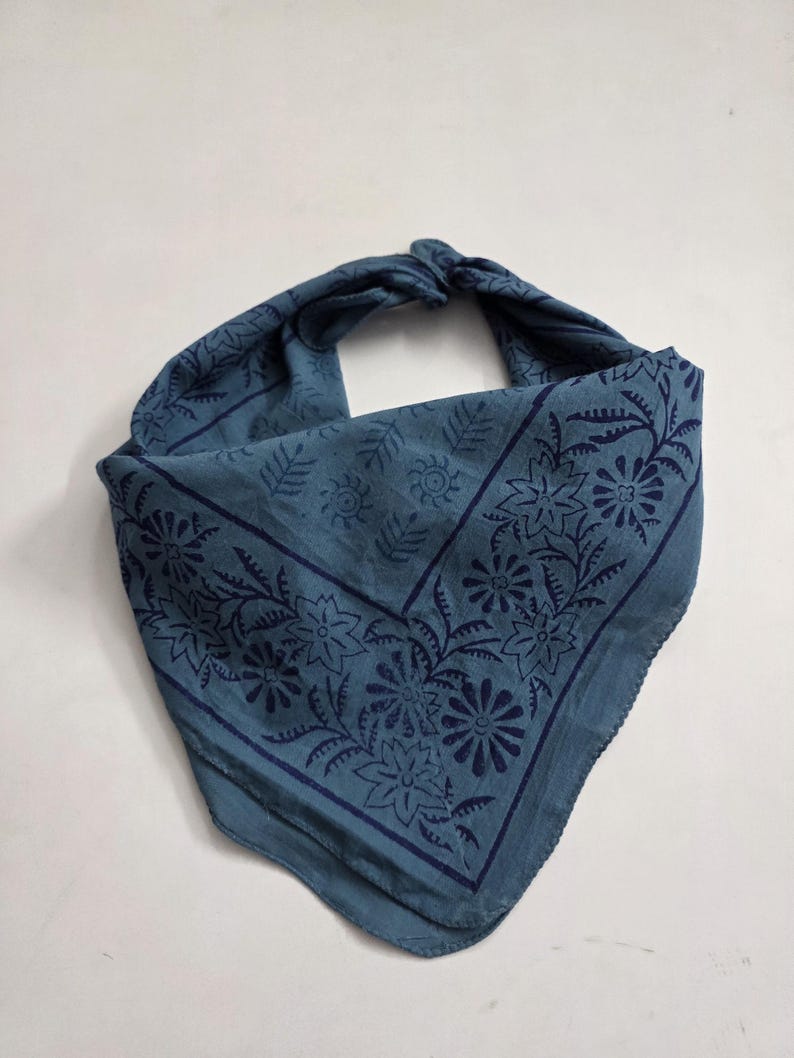 May include: A blue bandana with a floral pattern. The bandana is folded and tied at the top, showcasing the intricate dark blue floral design. The fabric has a soft, slightly wrinkled texture.