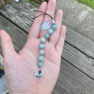 May include: A phone charm featuring a series of light blue round beads separated by small silver-toned spacers. The charm includes a translucent light blue leaf-shaped pendant and a small silver-toned bee charm with green accents at the end.