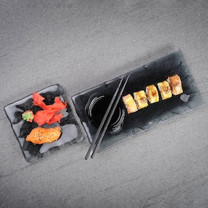 Puede incluir: Dos platos rectangulares negros con rollos de sushi. Un plato tiene salmón y jengibre, el otro tiene seis rollos con un lado de salsa de soja y palillos.