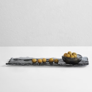 May include: A black glass serving tray with a wavy edge, featuring a small bowl filled with green olives. The tray is on a white surface.
