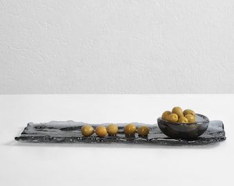 May include: A black glass serving tray with a wavy edge, featuring a small bowl filled with green olives. The tray is on a white surface.