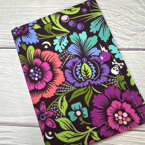 May include: A dark brown fabric wallet with a vibrant floral pattern in shades of pink, purple, blue, and green. The design includes spiderwebs, moons, and a skull and crossbones. It fastens with a purple button.