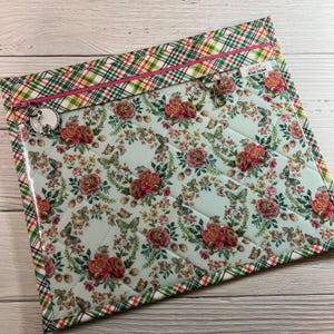 May include: A quilted zippered pouch with a clear vinyl front. The pouch has a plaid fabric exterior with a pink zipper and a floral print interior. The pouch has a small metal key ring attached to the zipper.
