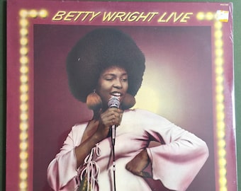 Op de afbeelding: Een vintage vinyl platenhoes met een vrouw met een afrokapsel die in een microfoon zingt. De platenhoes is rood met een gouden rand en de tekst "BETTY WRIGHT LIVE" in wit.