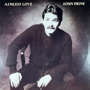 Puede incluir: Portada del álbum "Aimless Love" de John Prine. La imagen presenta a un hombre con bigote, vistiendo un suéter oscuro, sentado en una silla. El título del álbum y el nombre del artista están impresos en la parte superior de la portada.
