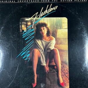 May include: Album cover for the "Flashdance" soundtrack. The image features a woman in a gray sweater and red heels. The title "Flashdance" is in a stylized script with blue accents. The top of the album reads "Original Soundtrack From The Motion Picture."